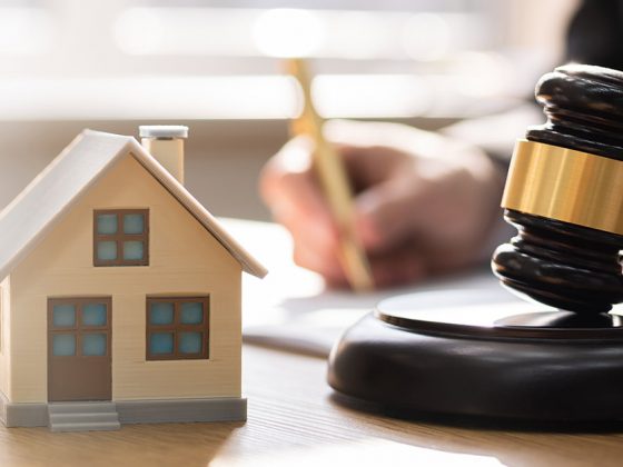 Ein Mann unterschreibt ein Dokument auf einem Tisch und im Vordergrund stehen ein Modellhaus und ein Richterhammer | Immobilie in der Scheidung