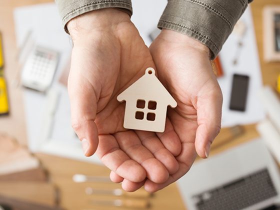 Real estate agent holding a small house, desktop with tools, wood swatches and computer on background, top view; Gebäudeenergie