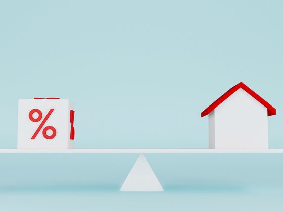 Vor einem hellblauen Hintergrund steht eine Waage, die ein Haus gegen Zinsen abwiegt | Jetzt Immobilie kaufen?
